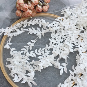Peut inclure: Garniture en dentelle blanche avec un motif de feuilles délicates. La garniture est disposée en cercle sur une surface grise.