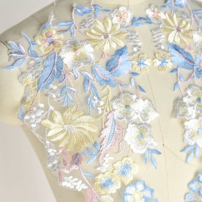 May include: A delicate floral lace applique with blue, yellow, white, and pink embroidery. The lace features a variety of floral designs, including small flowers, large flowers, and leaves.