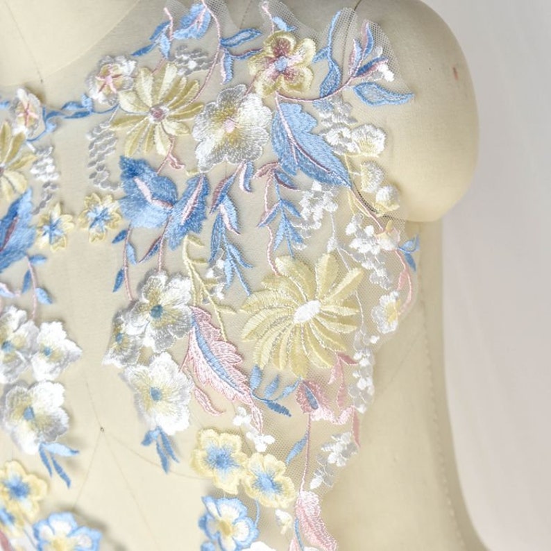 May include: A delicate floral lace applique with a light blue, yellow, and pink floral design. The lace is on a sheer mesh background.