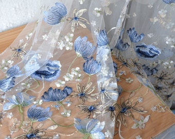 May include: Delicate white tulle fabric with embroidered blue and gold floral patterns. The design features large blue flowers, smaller white blossoms, and gold accents, creating an elegant and airy aesthetic. Ideal for bridal wear or decorative projects.