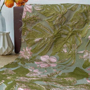 Op de afbeelding: Close-up van een groene stof met een bloemmotief. De stof heeft grote, getextureerde bladeren en roze bloemen met gouden accenten. De stof wordt waarschijnlijk gebruikt voor decoratieve doeleinden.