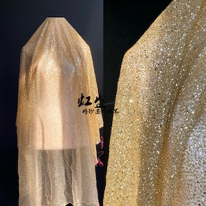 Puede incluir: Tela dorada con lentejuelas, drapeada. El tejido es de un color oro claro con una densa dispersión de pequeñas lentejuelas brillantes. El primer plano revela la textura y el brillo del material. La imagen también incluye texto en un idioma extranjero.