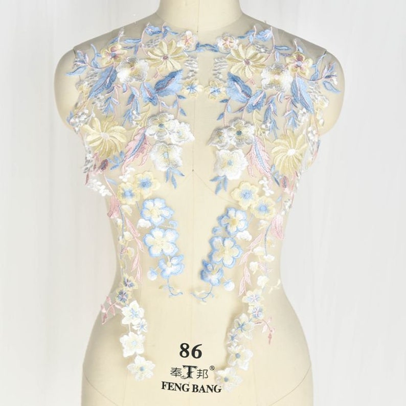 May include: A delicate floral lace applique featuring blue, yellow and pink flowers on a white mesh background. The lace is ideal for adding a touch of elegance to any project.