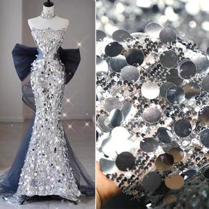 May include: A silver sequined evening gown on a mannequin. The strapless dress has a mermaid silhouette, a large navy blue bow, and a matching choker. The fabric is covered in large, round sequins. A close-up shows the sequin fabric.