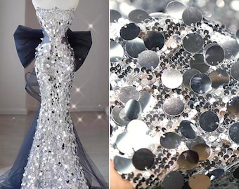 May include: A silver sequined evening gown on a mannequin. The strapless dress features a mermaid silhouette, a large navy blue bow, and a matching choker. The fabric is covered in large, round sequins. A close-up shows the sequin fabric.