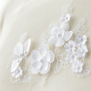 May include: White lace fabric with white fabric flowers and pearl accents. The flowers are arranged in a cluster and have a 3D effect.