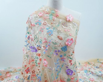 Op de afbeelding: Beige tule stof met kleurrijke geborduurde bloemen. De bloemen zijn in tinten roze, blauw, paars en groen.