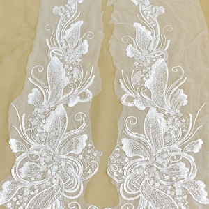 May include: Two white floral appliques with sequins and beads on a sheer white fabric background. The appliques are in the shape of a flower with a long stem and leaves.