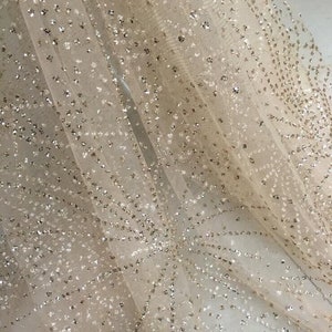 May include: Beige tulle fabric with a delicate pattern of silver glitter. The glitter is scattered throughout the fabric, creating a sparkling effect.
