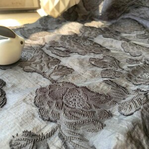 May include: A close-up of a gray fabric with a raised floral pattern. The fabric has a subtle sheen and a soft texture. A white ceramic rabbit figurine is sitting on the fabric.
