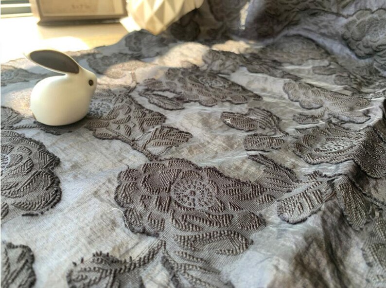 May include: A close-up of a gray fabric with a raised floral pattern. The fabric has a subtle sheen and a soft texture. A white ceramic rabbit figurine is sitting on the fabric.