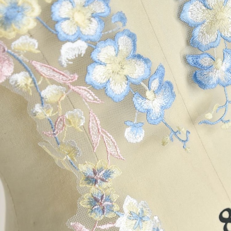 May include: Delicate floral embroidery featuring light blue, yellow, and pink flowers on a sheer white fabric. The embroidery is ideal for adding a touch of elegance to any project.
