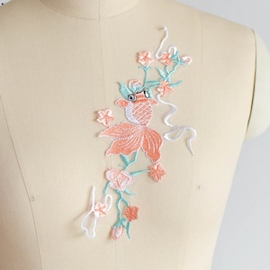 以下が含まれることがあります： 桃色とターコイズブルーの刺繍アップリケ。金魚と花のつるがデザインされています。