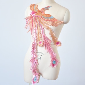 May include: A pink and orange embroidered phoenix applique with long, flowing feathers. The applique is on a white background.