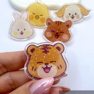 Könnte beinhalten: Eine Sammlung klarer Acryl-Anstecknadeln mit niedlichen Cartoon-Tiergesichtern: ein Hase, ein Küken, ein Hund, ein Bär und ein Tiger. Jede Anstecknadel hat einen glitzernden, holografischen Effekt und einen lächelnden Ausdruck. Die Anstecknadeln sind mit abgerundeten Kanten gestaltet.