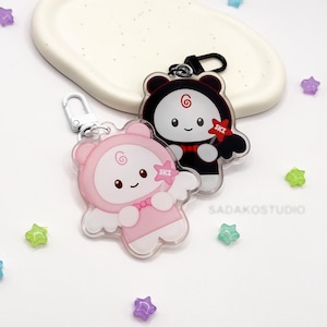 Puede incluir: Dos llaveros de acrílico con personajes de dibujos animados con orejas de oso y alas. Un personaje es rosa claro con cara blanca y el otro es negro con cara blanca. Ambos tienen una espiral roja en la frente y sostienen una estrella roja SKZ.