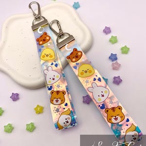 Könnte beinhalten: Zwei schillernde Schlüsselbänder mit Cartoon-Tierdesigns, darunter ein Tiger, ein Küken, ein Hase und ein Hund. Jedes Schlüsselband hat einen silbernen Verschluss und ist mit Stern- und Herzformen verziert. Die Schlüsselbänder liegen auf einem weißen Tablett mit sternförmigen Perlen.