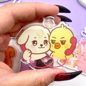 Könnte beinhalten: Transparenter Acryl-Schlüsselanhänger mit einem Cartoon-Hund und einem gelben Küken, die jeweils einen rosa Telefonhörer halten. Ein rosa herzförmiges Kabel verbindet die beiden Charaktere. Der Schlüsselanhänger hat einen kleinen herzförmigen Anhänger oben.