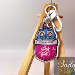 Può includere: Un portachiavi in acrilico trasparente con un personaggio di gatto blu con occhi gialli e un sorriso dentuto. Il gatto è all'interno di una tasca rosa con il testo "Sana Boys". Il portachiavi ha una chiusura color argento.