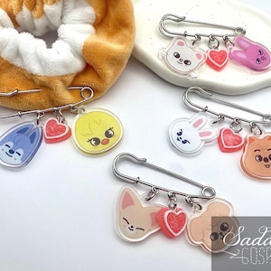 May include: A collection of clear acrylic pins with cartoon animal designs, including a cat, bunny, and chick, each paired with a red heart charm. The pins are attached to safety pins and are displayed on a white surface.