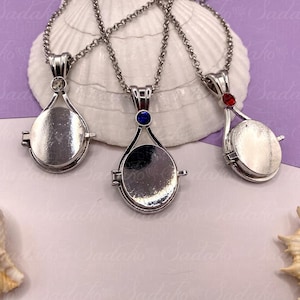May include: Three silver locket necklaces with teardrop pendants. Each pendant has a small gemstone at the top: clear, blue, and red. The lockets are on a silver chain, with a seashell and purple surface in the background.