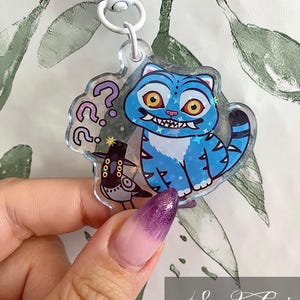 Pode incluir: Um chaveiro de acrílico transparente com um gato azul de desenho animado com olhos amarelos e um sorriso com dentes. O chaveiro também inclui um pequeno pássaro preto e pontos de interrogação. O chaveiro é preso a um fecho branco.