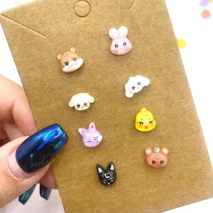 Può includere: Una collezione di piccoli orecchini a bottone colorati a forma di animali. Gli orecchini includono un criceto, un coniglio, un cane, un gatto, un pulcino, un orso e un coniglio viola. Sono esposti su una scheda marrone.