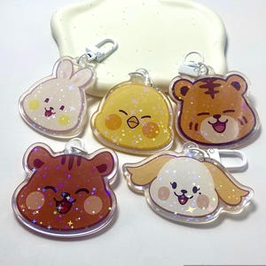 Könnte beinhalten: Fünf transparente Acryl-Schlüsselanhänger mit Cartoon-Tierdesigns: ein Hase, ein Küken, ein Tiger, ein Bär und ein Hund. Jeder Schlüsselanhänger hat einen silbernen Verschluss und ist mit Glitzer und lächelnden Gesichtern verziert. Die Schlüsselanhänger liegen auf einer weißen Oberfläche.