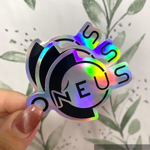 Könnte beinhalten: Holografische Aufkleber mit dem Wort "ONEUS" in einer stilisierten Schriftart. Die Aufkleber haben einen Regenbogen-Schimmer und eine schwarze Umrandung. Die Aufkleber werden in einer Hand vor einem Hintergrund mit einem Blättermuster gehalten.