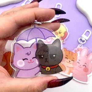 Peut inclure: Porte-clés en acrylique transparent représentant deux chats de dessin animé sous un parapluie violet. Un chat est violet et souriant, l'autre est noir avec un collier rouge et une clochette dorée. D'autres porte-clés avec des animaux de dessin animé sont visibles.
