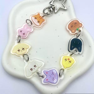 May include: A clear acrylic bracelet with cartoon animal faces, such as a tiger, bunny, and cat. The bracelet is linked with silver-coloured jump rings and a star-shaped clasp. The bracelet is on a white, heart-shaped dish.