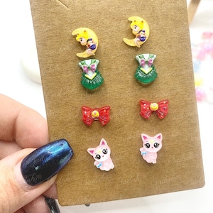 May include: A collection of six pairs of stud earrings presented on a brown card. The earrings showcase diverse designs: a yellow crescent moon with a figure, a green dress, a red bow, and a pink cat. These small, colourful earrings are ideal for everyday wear.