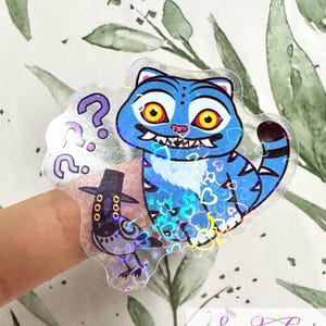 Könnte beinhalten: Ein holografischer Aufkleber mit einer blauen Cartoon-Katze mit gelben Augen und einem grinsenden Mund, sowie einer kleinen Vogel-Figur mit Hut. Fragezeichen befinden sich links. Der Aufkleber hat einen transparenten Hintergrund und den Text "Sadako COSPLAY".