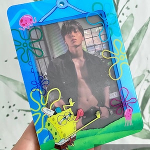 Può includere: Una cornice per foto in plastica blu con una foto di una persona. La cornice ha un tema di SpongeBob SquarePants con illustrazioni di SpongeBob, meduse e stelle marine. Il testo "Sadako COSPLAY" è visibile.