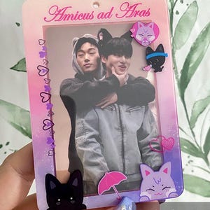 May include: A rectangular photo card with a gradient pink and purple background. The card features a photo of two people, cat-themed embellishments, and the text "Amicus ad Aras". Decorative elements include cat illustrations and a pink umbrella.