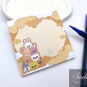 Könnte beinhalten: Quadratischer Haftzettel mit einer Cartoon-Illustration von gestapelten Tierfiguren in Braun-, Weiß-, Rosa- und Gelbtönen. Der Zettel hat einen wolkenartigen Rand und wird von zwei lila Stiften begleitet.