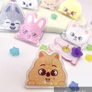 Puede incluir: Una colección de coloridos pines acrílicos de estilo caricatura con varios personajes de animales. Los pines incluyen un lobo, un conejo, un zorro, un pollito, un oso y un quokka, con detalles en forma de estrella. Los pines están sobre una superficie blanca.