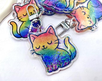 May include: Rainbow-colored cat keychain with a silver clasp. The cat design features a gradient of colors, including pink, yellow, green, and blue, with glitter accents. The cat is in a sitting position with its eyes closed.