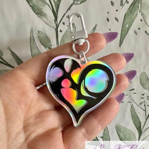 May include: A heart-shaped keychain with a black outline and a holographic, rainbow-colored center. The keychain has a silver clasp and a small ring. The design includes a circular shape and other abstract shapes. The keychain is held in a hand.