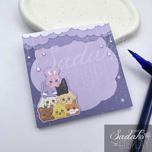 May include: A purple sticky note with a cloud-shaped design and a stack of cartoon animal stickers. The note has the text "Sada Cosplay" and is accompanied by a blue pen. The stickers include a bunny, cat, and other cute characters.