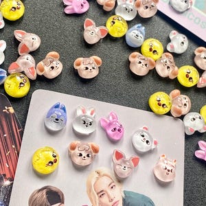 Könnte beinhalten: Eine Sammlung bunter Resin-Charms im Cartoon-Stil in verschiedenen Tierformen, darunter Hasen, Hunde und Katzen. Die Charms sind in den Farbtönen Rosa, Gelb, Blau und Transparent gehalten. Eine Fotokarte mit zwei Personen befindet sich im Vordergrund.