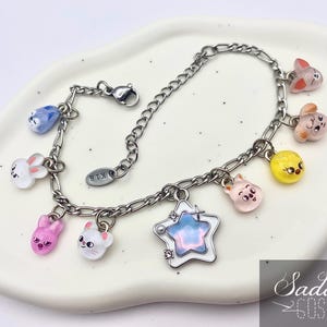 Könnte beinhalten: Silberfarbenes Charm-Armband mit bunten Cartoon-Anhängern. Zu den Anhängern gehören Hasen, Küken und ein Stern. Das Armband wird auf einem weißen, ovalen Teller präsentiert.