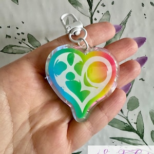 Könnte beinhalten: Ein herzförmiger Schlüsselanhänger mit Regenbogenverlauf und silbernem Verschluss. Der Schlüsselanhänger hat ein weißes Grafikdesign im Inneren der Herzform. Der Text "Sadako COSPLAY" ist unten rechts sichtbar.