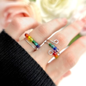 Könnte beinhalten: Zwei silberne Drahtringe, jeder mit einer Reihe farbiger Perlen in einem Regenbogenspektrum verziert. Die Ringe befinden sich an einem Finger, mit einem schwarzen Ärmel im Hintergrund. Die Perlen sind in Rot-, Orange-, Gelb-, Grün-, Blau- und Lilatönen gehalten.