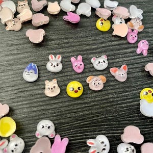 Könnte beinhalten: Eine Sammlung bunter, kleiner Dekorationsperlen in verschiedenen Formen, darunter Tiere wie Hasen, Bären und Küken. Die Perlen sind in den Farbtönen Rosa, Gelb, Weiß und Beige gehalten und auf einer dunklen Oberfläche verstreut.