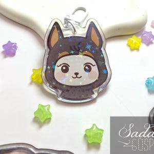 Puede incluir: Un dije acrílico transparente con un personaje de perro de dibujos animados con un atuendo con capucha marrón oscuro con estrellas. El dije tiene un cierre plateado y está rodeado de cuentas en forma de estrella de colores.