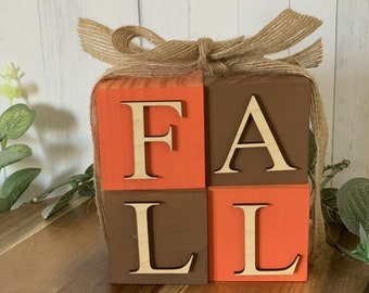 May include: Wooden block letters spelling "FALL" in orange and brown paint. The blocks are tied together with twine.