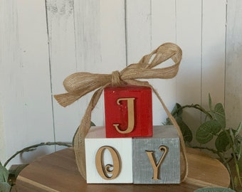 May include: Wooden blocks spelling "JOY" in gold letters. The blocks are stacked on top of each other, with the red block on top, the white block in the middle, and the gray block on the bottom. The blocks are tied together with a brown ribbon.