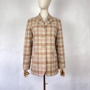 May include: A tan and pink plaid blazer with three buttons and two front pockets. The blazer is displayed on a mannequin and has a classic, tailored design. The plaid pattern is a mix of tan, pink, and cream colors.
