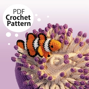May include: PDF Crochet Pattern for a crocheted orange and white clownfish with black stripes, sitting on a crocheted purple and cream anemone.